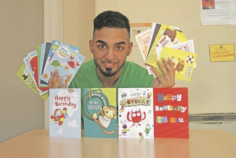 A man sat behind a table displaying birthday cards and posing with birthday cards in his hands.